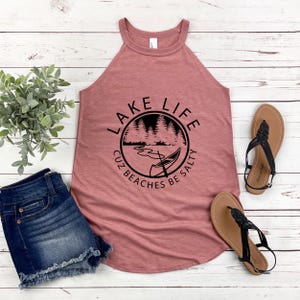 May include: A dusty rose tank top with a black graphic that reads "Lake Life" with a lake scene. The tank top is paired with denim shorts and brown sandals with black straps. A small green plant is also in the image.