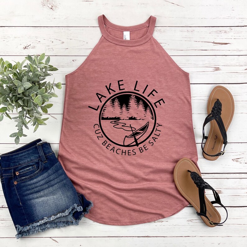 May include: A dusty rose tank top with a black graphic that reads "Lake Life" with a lake scene. The tank top is paired with denim shorts and brown sandals with black straps. A small green plant is also in the image.