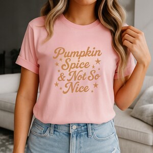 May include: A light pink short-sleeved t-shirt featuring the brown text Pumpkin Spice & Not So Nice printed on the front. The design includes several small star icons scattered around the lettering, which is written in a cursive script font.
