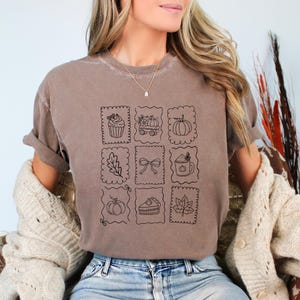 May include: A brown short-sleeve t-shirt featuring a grid of nine black line-art illustrations. The designs include a cupcake, a wagon with pumpkins, a pumpkin, an oak leaf, a bow, a mug, a round pumpkin, a slice of pie, and a maple leaf.