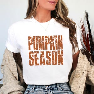 May include: A white short-sleeved t-shirt featuring the text PUMPKIN SEASON printed in a distressed, burnt orange font across the chest. The shirt has a crew neckline and is worn with a beige knit cardigan and blue denim jeans.