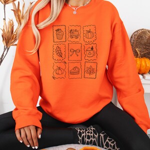 May include: A bright orange long-sleeve sweatshirt featuring a black line art graphic print. The design consists of nine individual square frames containing illustrations of a cupcake, a wagon with pumpkins, a pumpkin, a leaf, a bow, a mug, a pumpkin, a slice of pie, and a maple leaf.