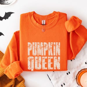 May include: A bright orange long-sleeve crewneck sweatshirt featuring the text PUMPKIN QUEEN in a distressed, off-white block font centered on the chest. The garment has a ribbed neckline and cuffs, with a silver-toned sunburst pendant necklace.