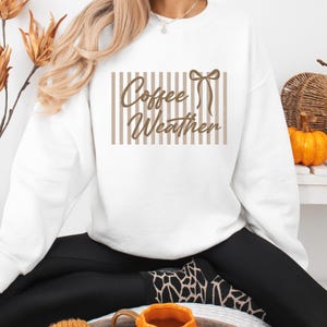 May include: A white long-sleeve crewneck sweatshirt featuring a brown graphic print on the chest. The design consists of vertical stripes, a bow icon, and the brown cursive text that reads Coffee Weather centered within the striped rectangular area.