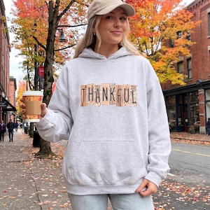 May include: A light gray hooded sweatshirt featuring the word THANKFUL printed in black capital letters across the chest. Each letter is set against a rectangular background with various autumn-themed patterns including plaid, leaves, and wood grain textures.