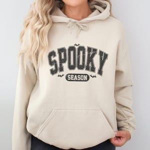May include: A beige hooded sweatshirt featuring a distressed black graphic on the chest that reads SPOOKY SEASON in a bold collegiate font. The design includes small bat silhouettes above and below the text, with a front kangaroo pocket.