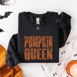 May include: A black long-sleeve sweatshirt featuring the text PUMPKIN QUEEN in a distressed orange font across the chest. The garment has a crew neckline and a white brand label inside the collar. A silver sunburst pendant necklace is visible.