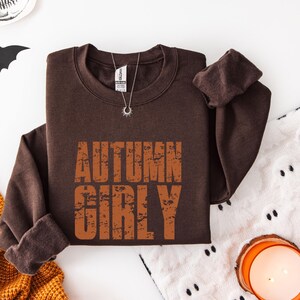 May include: A dark brown crewneck sweatshirt featuring the text AUTUMN GIRLY in a distressed orange font. The garment has long sleeves, a ribbed collar, and a silver-toned necklace with a sunburst pendant resting on the neckline area.