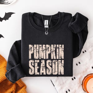 May include: A black crewneck sweatshirt featuring the text PUMPKIN SEASON in a distressed, light beige font across the chest. The garment has long sleeves with ribbed cuffs and a ribbed neckline, with a silver sunburst pendant necklace resting on it.