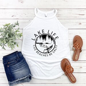 May include: White tank top with a black graphic that reads "Lake Life Cuz Beaches Be Salty." The image also includes denim shorts and brown sandals. The tank top has a halter neckline.