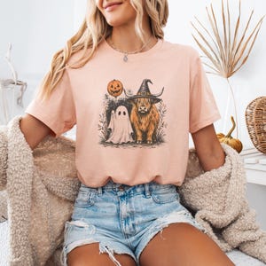 May include: A light peach-colored short-sleeve t-shirt featuring a graphic print of a Highland cow wearing a witch hat, standing next to a ghost holding a jack-o'-lantern balloon. The design includes subtle botanical elements and a textured ink style.