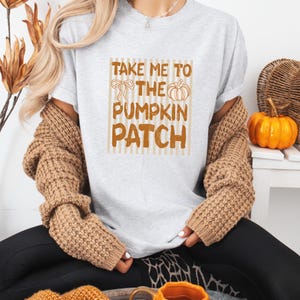 May include: A light gray short-sleeve t-shirt featuring a brown graphic print with vertical stripes, a bow, a pumpkin, and the text "TAKE ME TO THE PUMPKIN PATCH". The shirt is worn under a tan textured long-sleeve cardigan with a chunky knit pattern.