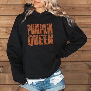 May include: A black hooded sweatshirt featuring the text PUMPKIN QUEEN in a distressed orange font on the chest. The garment includes a front kangaroo pocket, long sleeves, and a hood with drawstrings, paired with light blue denim shorts.