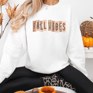 May include: A white long-sleeve crewneck sweatshirt featuring the text FALL VIBES printed in black capital letters. Each letter is contained within a vertical rectangular block decorated with various orange, brown, and white autumn-themed patterns.