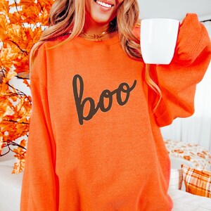 May include: A bright orange long-sleeve sweatshirt featuring the word boo written in a dark brown cursive script across the chest. The garment has a crew neckline and long sleeves, providing a solid color base for the dark brown text graphic.