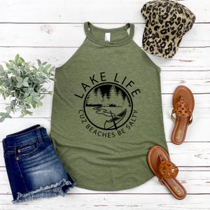 May include: Olive green tank top with the text "LAKE LIFE cuz beaches be salty" in a black circular design. The image also includes denim shorts, brown sandals, and a leopard print baseball cap.