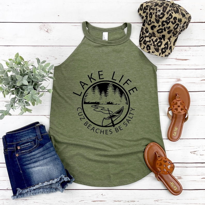 May include: Olive green tank top with the text "LAKE LIFE cuz beaches be salty" in a black circular design. The image also includes denim shorts, brown sandals, and a leopard print baseball cap.