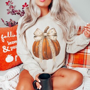 May include: A beige long-sleeve sweatshirt featuring a central graphic of an orange pumpkin adorned with a large, decorative cream-colored bow. The pumpkin design includes small star accents and lace-like trim details on the ribbon streamers.