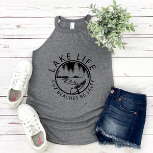 May include: A gray tank top with the words "LAKE LIFE" in a circular design with a lake and canoe graphic. The text "CUZ BEACHES BE SALTY" is below the graphic. The tank top is paired with denim shorts and white sneakers.