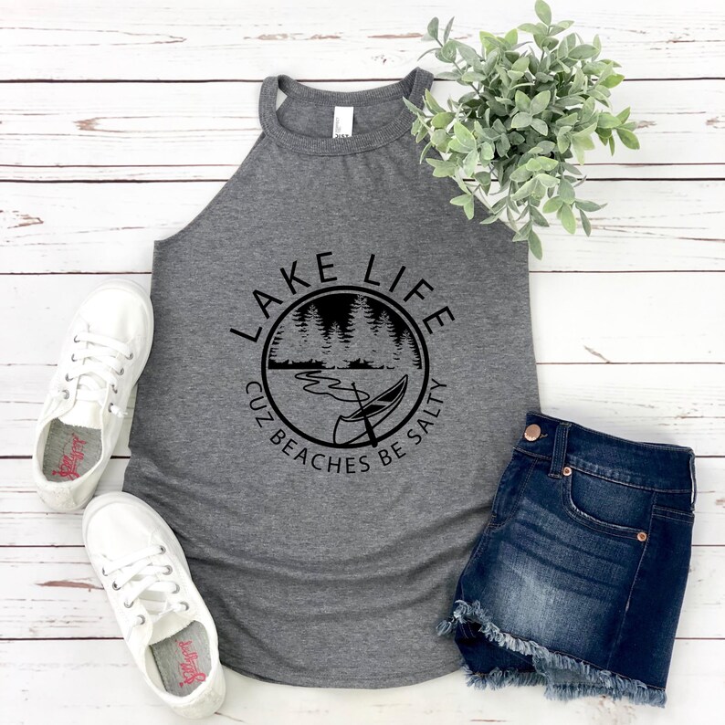 May include: A gray tank top with the words "LAKE LIFE" in a circular design with a lake and canoe graphic. The text "CUZ BEACHES BE SALTY" is below the graphic. The tank top is paired with denim shorts and white sneakers.
