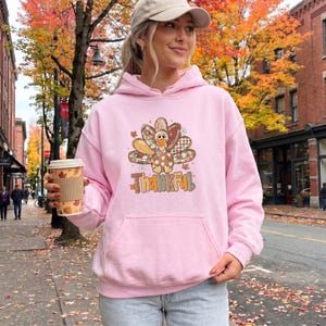 May include: A light pink hooded sweatshirt featuring a graphic of a turkey with patterned feathers in shades of brown, orange, and white. The word Thankful is printed below the turkey in a decorative font. The hoodie has a front kangaroo pocket.