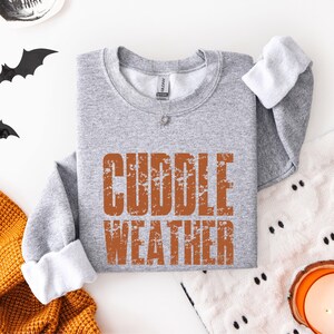 May include: A heather gray crewneck sweatshirt featuring the text CUDDLE WEATHER printed in a distressed, burnt orange font across the chest. The garment has long sleeves with ribbed cuffs and a standard neckline with a visible brand label.
