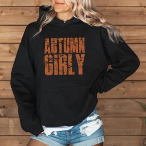 May include: A black hooded sweatshirt featuring the text AUTUMN GIRLY printed in a distressed, burnt orange font across the chest. The garment includes a front kangaroo pocket, long sleeves, and a hood with matching black drawstrings.