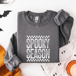 May include: A dark gray crewneck sweatshirt featuring the white text "SPOOKY SEASON" in a distressed font, centered between two rows of white checkered patterns. The garment has long sleeves, a ribbed collar, and a silver-toned sunburst pendant necklace.