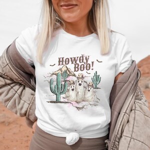 May include: A white t-shirt featuring a graphic print with the text Howdy Boo! above three ghosts wearing cowboy hats, surrounded by green cacti, a desert mountain landscape, and small black bats, with pink and brown watercolor accents.