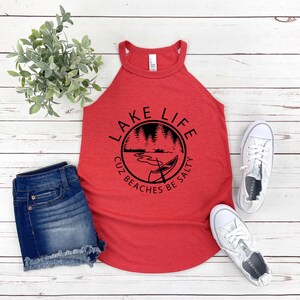 May include: Red tank top with the text "LAKE LIFE CUZ BEACHES BE SALTY" in a black circular design. The design includes a lake scene with a canoe. The tank top is paired with denim shorts and white sneakers.
