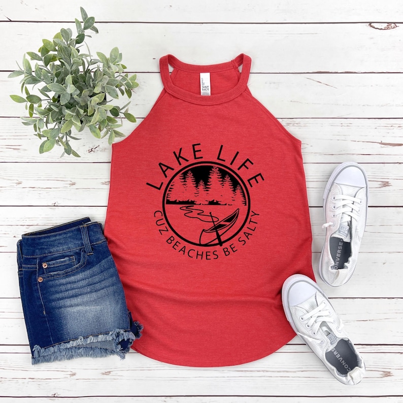 May include: Red tank top with the text "LAKE LIFE CUZ BEACHES BE SALTY" in a black circular design. The design includes a lake scene with a canoe. The tank top is paired with denim shorts and white sneakers.
