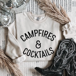 May include: Cream-colored sweatshirt with the text "Campfires & Cocktails" in black. The sweatshirt is paired with a black cowboy boot with white stitching. Decorative elements include a disco ball and dried floral accents.