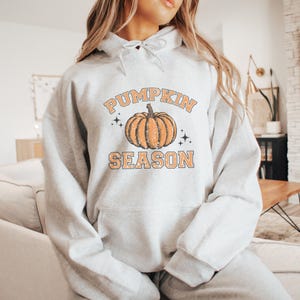 May include: A light gray hooded sweatshirt featuring a graphic print of an orange pumpkin centered between the words PUMPKIN and SEASON in a distressed orange font. The design includes small black star accents and a front kangaroo pocket.