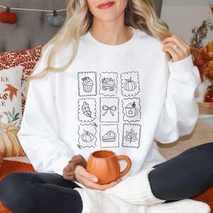 May include: A white long-sleeve sweatshirt featuring a central graphic print of nine rectangular postage-stamp-style frames. Each frame contains a black line drawing of autumn-themed items including a cupcake, pumpkin, leaf, bow, and pie.