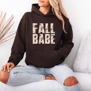 May include: A dark brown hooded sweatshirt featuring a front kangaroo pocket and the text FALL BABE printed in a distressed, light beige font across the chest. The garment has long sleeves and a hood with matching drawstrings at the neckline.