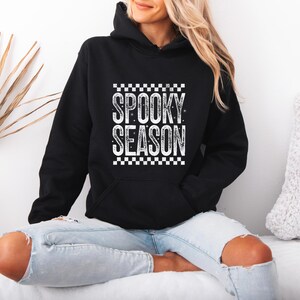 May include: A black hooded sweatshirt featuring a white graphic print on the chest. The design includes the text SPOOKY SEASON in a distressed font, framed by a checkered pattern at the top and bottom, with small star accents throughout the lettering.