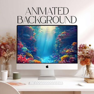 Op de afbeelding: Een zilveren desktopcomputermonitor toont een onderwaterkoraalrifscène met levendige oranje, gele en rode koraalformaties. De tekst "ANIMATED BACKGROUND" staat in het zwart boven het scherm. Een wit toetsenbord ligt op het bureau.