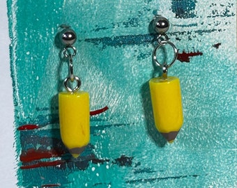 May include: A pair of dangle earrings featuring yellow pencil-shaped charms with gray tips. The earrings hang from silver-colored ball studs. The earrings are displayed on a white card with a teal and red painted design.