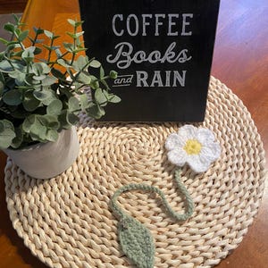 May include: A crochet bookmark shaped like a white daisy with a yellow centre and a green leaf. The bookmark rests on a woven placemat, next to a black sign that reads "COFFEE Books and RAIN" and a small potted plant.