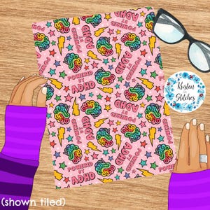 May include: Pink fabric featuring a repeating pattern of colourful brains, stars, and lightning bolts. The text "POWERED BY ADHD" is also featured. A pair of glasses and a sticker are in the background.