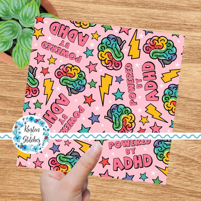 May include: A pink square featuring a repeating pattern of colourful brains, stars, and lightning bolts. The words "POWERED BY ADHD" are also present. The design is held by a hand, with a plant in the background.