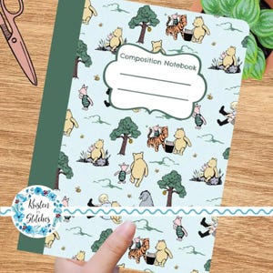 K&ouml;nnte beinhalten: Ein hellblaues Notizbuch mit Illustrationen von Winnie Puuh und seinen Freunden. Der Einband tr&auml;gt den Text "Composition Notebook" in einem wei&szlig;en Etikett. Das Notizbuch hat einen gr&uuml;nen R&uuml;cken und wird von einer Hand gehalten.