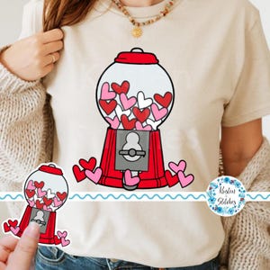 Op de afbeelding: Beige T-shirt met een rode kauwgomballenautomaat-afbeelding gevuld met rode, roze en witte harten. De kauwgomballenautomaat heeft een rode basis en bovenkant, met een zilveren muntsleuf. Verschillende hartvormen zijn rond de machine verspreid.