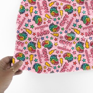 May include: Pink fabric featuring a repeating pattern of colourful brains, stars, and lightning bolts. The text "POWERED BY ADHD" is printed across the design. The fabric is held by a hand with gold rings.