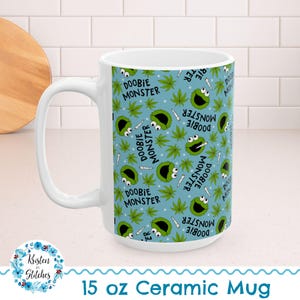 May include: White 15 oz ceramic mug with a repeating pattern of a green monster character, marijuana leaves, and cigarettes on a light blue background. The mug has the text "DOOBIE MONSTER".