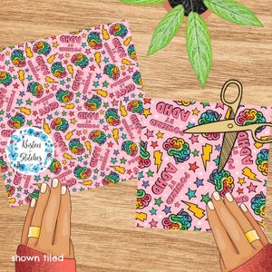 May include: Two pink fabric pieces with a repeating pattern of colourful brains, stars, and lightning bolts, with the text "POWERED BY ADHD". A pair of gold scissors cuts one piece. A plant is in the background.