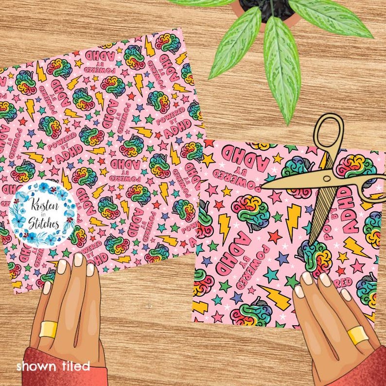 May include: Two pink fabric pieces with a repeating pattern of colourful brains, stars, and lightning bolts, with the text "POWERED BY ADHD". A pair of gold scissors cuts one piece. A plant is in the background.