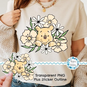 May include: Beige t-shirt featuring a cartoon bear character surrounded by cream and white flowers with green leaves. The text "Transparent PNG Plus Sticker Outline" is printed at the bottom. A sticker with the text "Kristen Stitches" is on the right.