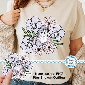 Op de afbeelding: Beige T-shirt met een cartoon ezel ontwerp, omringd door lichtpaarse en roze bloemen en groene bladeren. De tekst "Transparent PNG Plus Sticker Outline" staat onderaan gedrukt. Een sticker met de tekst "Kristen Stitches" staat rechts.