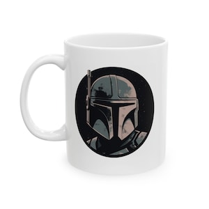 May include: White ceramic mug featuring a stylized illustration of a Mandalorian helmet within a black circle. The helmet is depicted in shades of gray and tan, set against a starry background. The mug has a curved handle.